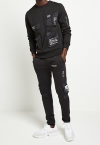 Černá mikina a tepláky z měkkého materiálu, s několika texturovanými kapsami s bílými potištěnými štítky. Oblečení doplňují bílé tenisky.