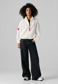 Λευκό τζάκετ από φλίς με κίτρινο φερμουάρ και κόκκινη ετικέτα, συνδυασμένο με μαύρα τζιν με φαρδιές παντελόνι και γκρι αθλητικά παπούτσια. Υφασμάτινη υφή και άνετη εφαρμογή.