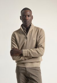 Suéter beige con cuello de cremallera a cuarto, tejido texturizado, puños acanalados y un pequeño logo en el pecho, combinado con pantalones marrón claro a juego.