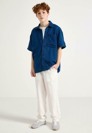 Ung person med krøllet rødt hår iført en løs blå kortærmet skjorte, hvid T-shirt, hvide bukser og grå sneakers, stående foran en ensfarvet baggrund.