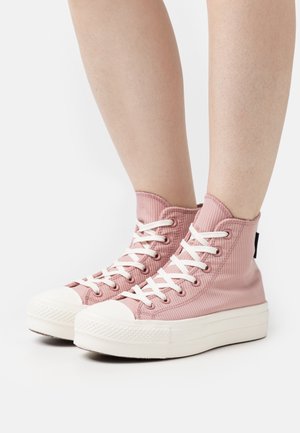 Personne portant des baskets montantes roses avec des lacets blancs et des semelles blanches épaisses, debout sur un fond blanc uni.