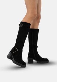 Botas negras de ante hasta la rodilla con correa en el tobillo, herrajes plateados, suelas texturizadas y tacones apilados. Cierre lateral con cremallera y detalle de hebilla ajustable.