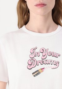 Tricou alb din bumbac, cu un text grafic roz "În visele tale" și o ilustrație cu ruj, cu accente roz și contururi negre.
