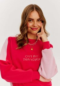 Červeno-růžová mikina s širokými rukávy, na jejímž středu je bíly text "CUTE BUT OVERTHINKER" a má měkkou texturu.