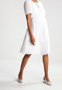 Valkoinen mekko, jossa on lyhyet hihat, solmimisyksityiskohta kaula-aukossa ja kirjailtu helmareuna. Yhdistetty hopeisiin korkokenkiin.