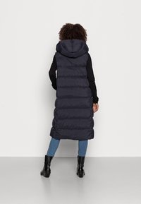 Marinblå vadderad väst med huva, som har lång längd, kviltat mönster och slät textur. Är matchad med skinny jeans och ankelstövlar.