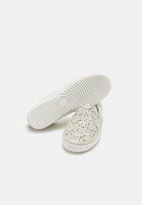 Kangan slip-on berwarna beige terang yang menampilkan desain ter perforasi, sol karet bertekstur, dan detail jahitan putih. Ujung bulat dan bodi yang fleksibel.