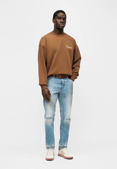 Άνδρας που στέκεται με τα χέρια στις τσέπες, φοράει καφέ φούτερ, ανοιχτό μπλε τζιν με σκισίματα και λευκά αθλητικά παπούτσια σε απλό φόντο.