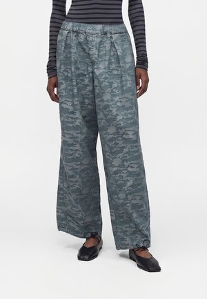 Žena má na sebe šedé maskáčové nohavice s širokými nohavicami a elastickým pásom, v kombinácii s čiernymi plochými topánkami so štvorcovou špičkou a čierno-šedo pruhovaným dlhým rukávom.