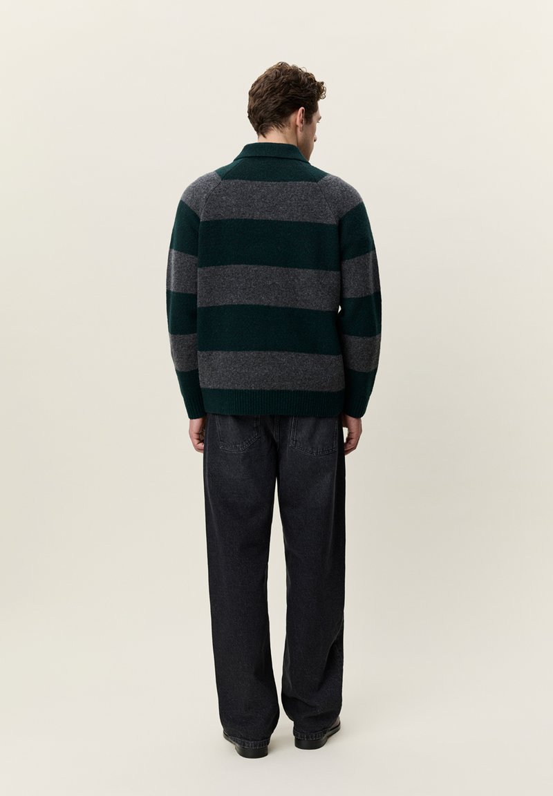 Homme portant un pull à larges rayures vert foncé et gris avec un jean large noir, debout de dos sur un fond clair uni.