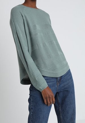 Hellgrüner Strukturpullover mit Boot-Ausschnitt, langen Ärmeln und lockerer Passform, kombiniert mit blauen Jeans.