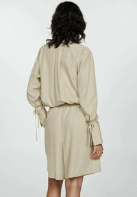 Beige short dress with long sleeves, featuring tie details at the cuffs and an elastic waist for a relaxed fit. Soft, smooth fabric.
