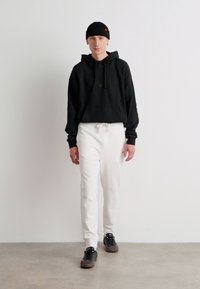 Sort hættetrøje med kangaroo-lomme, kombineret med hvide bukser. Modellen har en sort beanie og mørke sneakers på. Glat tekstur.