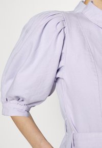 Vestido camiseiro lilás com mangas bufantes, gola e uma cintura definida com detalhes de cinto. Feito de um tecido leve e macio.