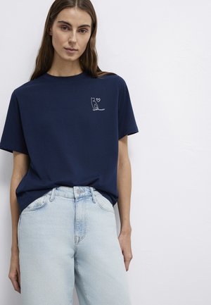 Woman wearing a navy blue T-shirt with a small white cat and heart design on the chest, paired with light blue high-waisted jeans.