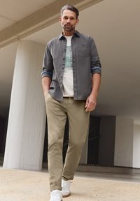 Homme barbu portant une chemise grise à boutons, un t-shirt blanc imprimé, un pantalon beige et des baskets blanches, marchant sous une structure en béton.