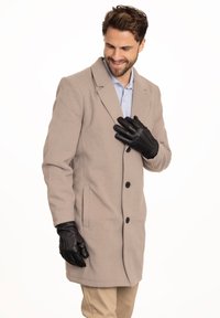 Beiger Wollmantel mit Reverskragen, Knopfverschluss und seitlichen Taschen. Trägt schwarze Lederhandschuhe mit feinen Nähdetails.