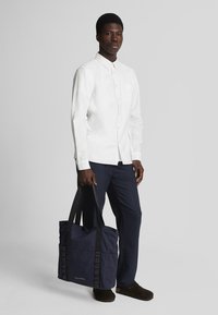 Marineblauwe tote bag met zwarte handvatten, met een gladde textuur en een minimalistisch ontwerp. De tas heeft verticale webbingdetails aan de zijkanten.