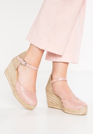 Fødder iført lyserøde espadrille kilehæl med ankelremme, kombineret med lyserøde cropped bukser på hvid baggrund.