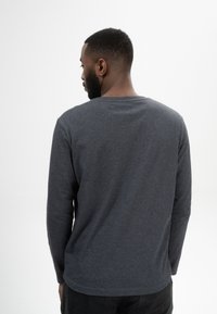 Mann mit kurzem Haar und Bart, der ein langärmeliges, dunkelgraues Hemd trägt und mit dem Rücken zu einem schlichten weißen Hintergrund steht.