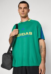 Zöld, túlságosan nagy póló sárga és kék díszítéssel, az elején "ADIDAS SPORTSWEAR" felirattal. Fekete táska lóg a vállon.