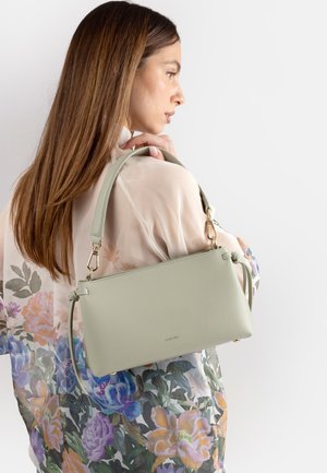 Donna con lunghi capelli castani che indossa una camicetta trasparente a fiori tiene una borsa a tracolla in pelle verde chiaro su uno sfondo uniforme.