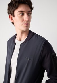 Στρατιωτική βομβιστική ζακέτα σε ναυτικό χρώμα με ομαλή μπροστινή επιφάνεια, ριγωτό γιακά και επενδεδυμένη τσέπη στο μανίκι. Ξεχωριστό λογότυπο "BOSS" κεντημένο στο στήθος.