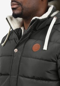 Fekete steppelt dzseki fehér műszőrmével bélelve, elöl gombokkal és bőr logófolttal. Kapucnival és húzózsinóros részletekkel.