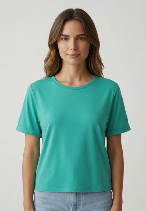 Giovane donna che indossa una t-shirt tinta unita color teal a maniche corte con scollo tondo e jeans azzurri, in piedi davanti a uno sfondo grigio neutro.