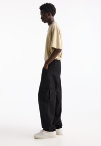 T-shirt beige oversize, pantaloni cargo neri con molte tasche e sneakers bianche. L'outfit presenta una vestibilità comoda e una consistenza morbida.