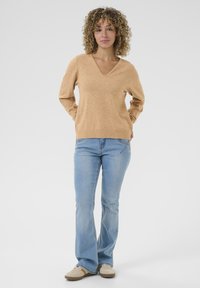 Beige v-halstrui, gebreid textuur, gecombineerd met lichtblauwe uitlopende jeans. Model draagt beige sneakers; handen op de heupen. Eenvoudige, casual outfit.