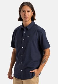 Camisa de botones de manga corta en color navy, confeccionada en una tela texturizada, que presenta un pequeño logo blanco en el pecho y botones blancos clásicos.