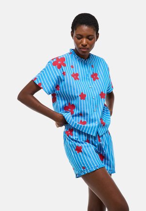 Femme portant une chemise bleue à manches courtes boutonnée et un short assorti avec des rayures blanches et des motifs de fleurs rouges, debout, les mains sur les hanches.