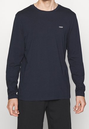 Mann trägt ein marineblaues Langarmshirt mit kleinem weißen "HUGO"-Logo auf der Brust, kombiniert mit schwarzen Hosen, steht vor einfarbigem Hintergrund.