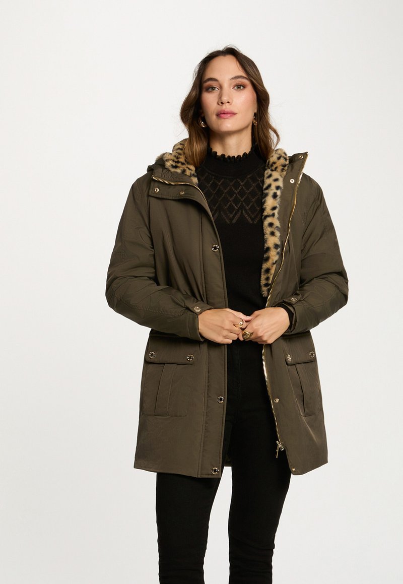 Donna che indossa un parka verde oliva con un sottopelo in stampa leopardata, un maglione nero a fantasia e pantaloni neri su uno sfondo chiaro e semplice.