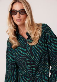Czarna koszula z długim rękawem zapinana na guziki, z falistymi paskami w zielonym i turkusowym kolorze; wykonana z lekkiego materiału; w połączeniu z czarnymi okularami przeciwsłonecznymi w owalnym kształcie.
