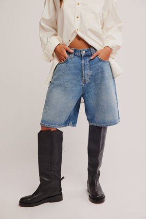 Kéz a laza, kék farmer rövidnadrág zsebében, melyet magas fekete bőr csizmával és törtfehér gombos inggel párosítanak.