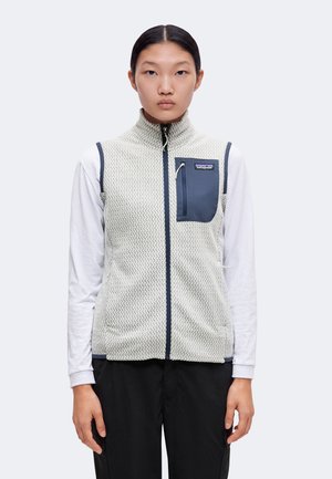 Young woman wearing light gray patterned zip-up vest with dark pocket over white long sleeve shirt and black pants, standing against plain background.