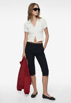 Frau mit schwarzen Sonnenbrillen, trägt ein weißes, kurzärmeliges, geknöpftes Oberteil, schwarze Kniebundhose, hält eine rote Jacke und trägt schwarze, gepunktete Ballerinas.