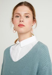 Young woman with light brown hair wearing translucent peach hoop earrings, white collared shirt, and blue knit sweater against a plain background.
