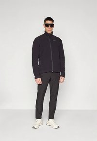 Vyras, vilkintis juodą zip-up gyvenimo striukę, juodus kelnes, didelius juodus akinius nuo saulės ir baltus sportinius batus, stovi prieš neutralų foną.