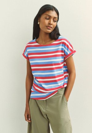 Kvinde med langt mørkt hår iført guldringeøreringe, en kortærmet skjorte med røde, hvide og blå striber og olivengrønne bukser.