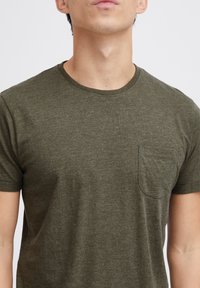 T-shirt olive vert à manches courtes en tissu doux, avec un col rond et une petite poche poitrine. Texture globale lisse.