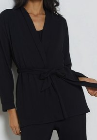 Giacca avvolgente nera a coste con maniche lunghe, vita con cintura e scollatura a V profonda. Texture morbida con vestibilità ampia, ideale per essere indossata a strati.