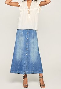 Lichte witte blouse met ruches aan de mouwen, gecombineerd met een lange denimrok met knopen aan de voorkant en een lichte wassing. Sleehakken met banden.