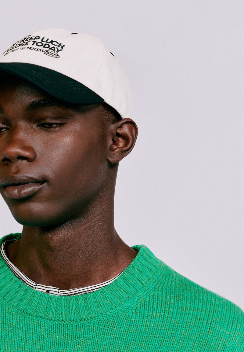 Jeune homme à la peau foncée portant une casquette blanche avec un texte et un pull en maille vert par-dessus une chemise à rayures sur un fond uni.