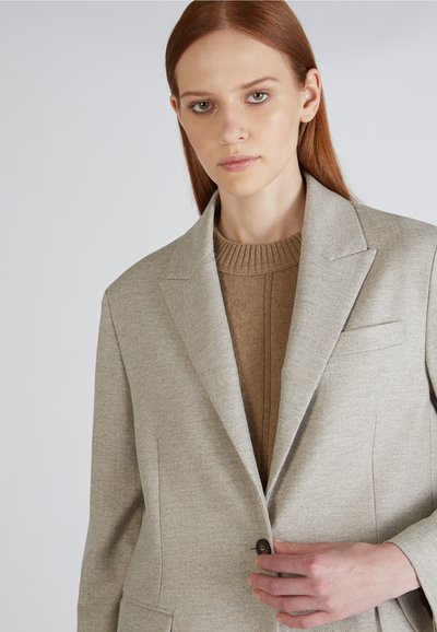 Blazer beige a spina di pesce con rever a lobon, un solo bottone e due tasche frontali, indossato sopra un maglione in maglia marrone texturizzato.