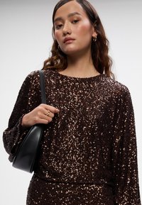 Smeđa bluzica od šljokica s dugim rukavima, okruglim izrezom i rebrastim donjim dijelom. Model drži malu crnu torbicu i nosi prsten. Glatka tekstura.