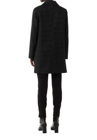 Black textured coat with a collar, mid-length, featuring a single button closure, paired with black fitted pants and ankle boots.