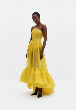 Donna che indossa un abito lungo giallo acceso senza spalline con orlo arricciato, in piedi con le mani vicino ai fianchi, su sfondo bianco uniforme.